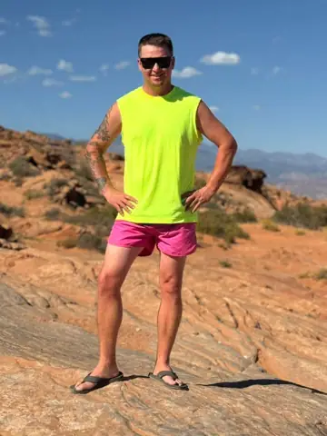 Brett in a neon yellow tank top and pink shorts, posing on red rocks